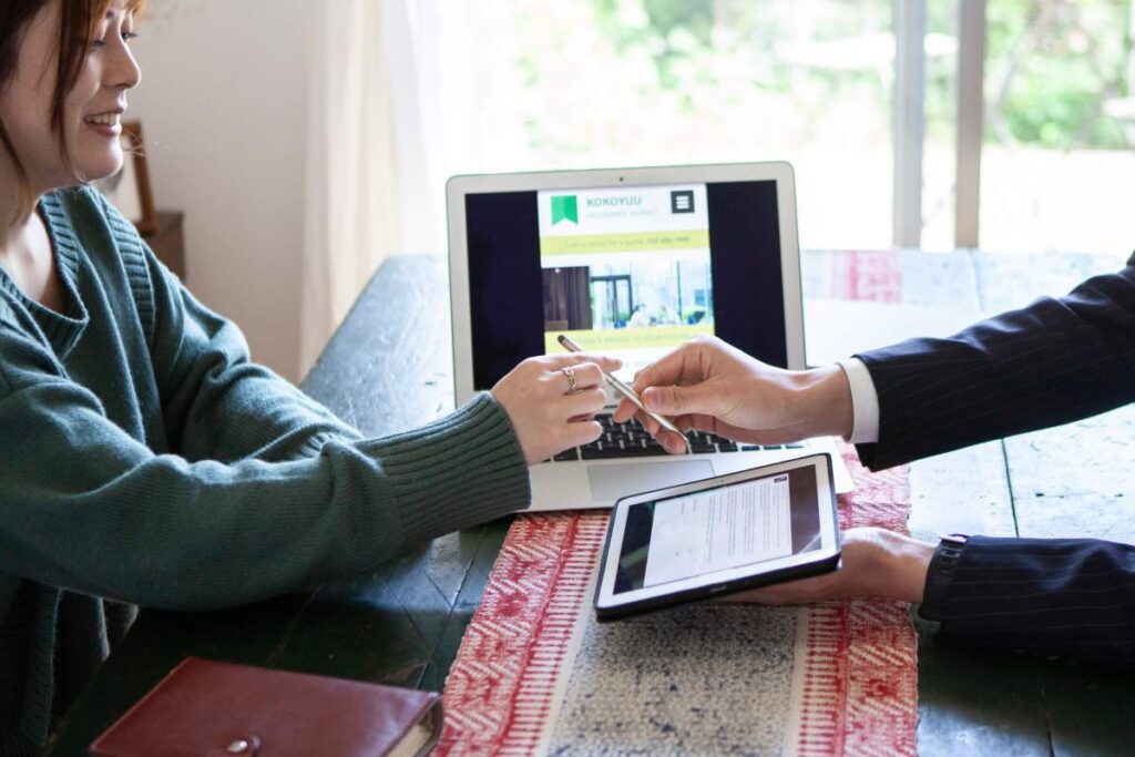 A diverse group of adults discusses community updates around a table with laptops and printed materials.